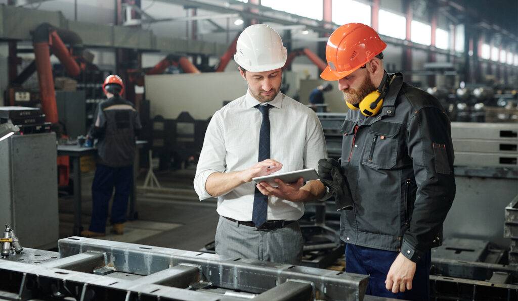 Two men in a factory setting wearing hard hats reviewing information on a tablet