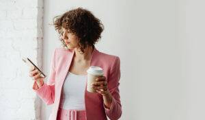 Woman wearing a pink business suit holding a coffee and looking at her phone as if waiting for a message