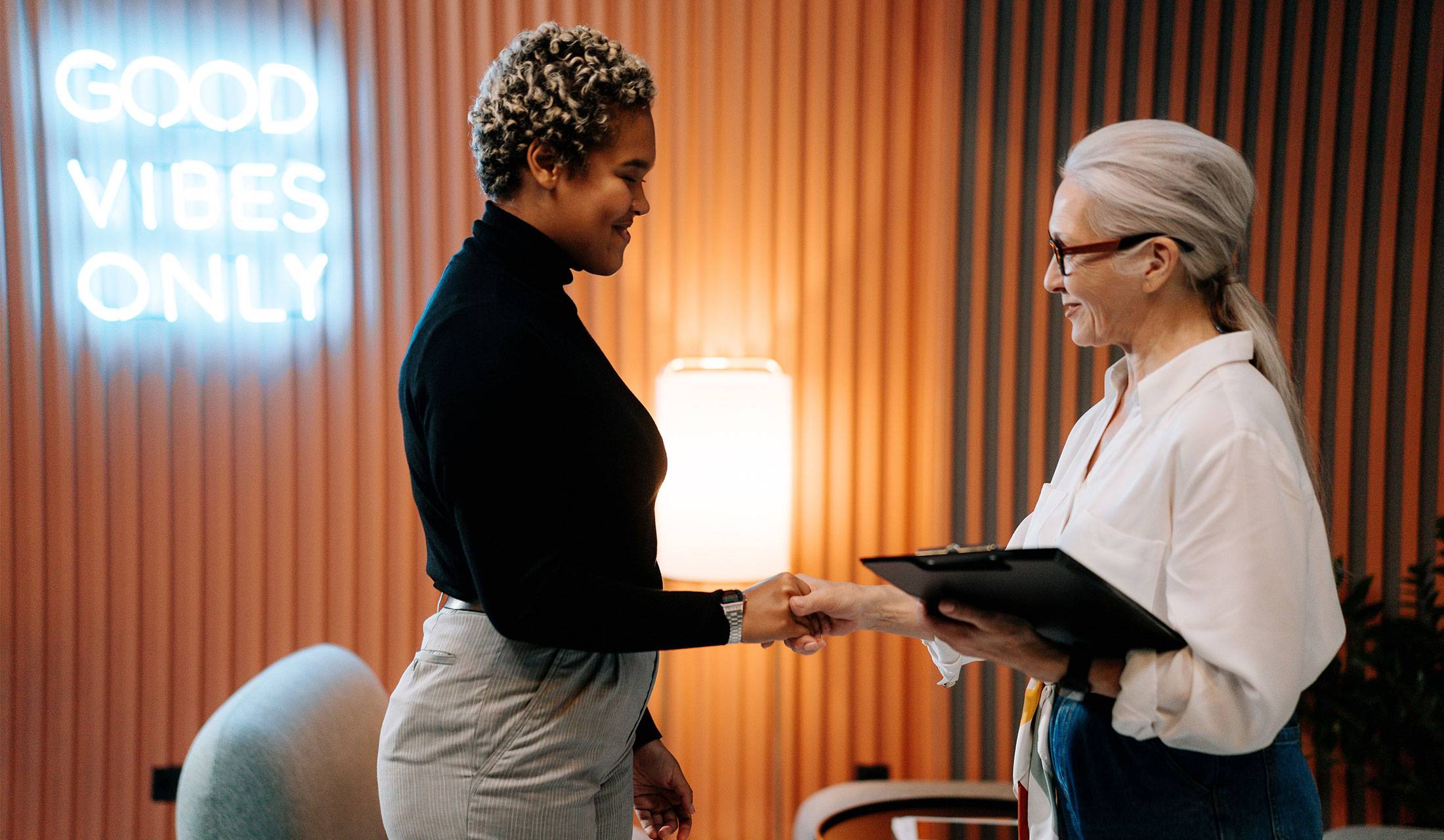 A manager shaking hands with a new employee in an office, with a neon sign in the background that reads Good Vibes Only